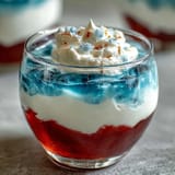 Red, white, and blue layered Jello cups in clear glasses, showing vibrant stripes of strawberry, creamy white, and blue raspberry gelatin for a festive look.